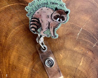 May include: A raccoon-themed badge reel with the text "I got 99 problems and I caused them all." The raccoon is brown and tan with a black mask and striped tail. The badge reel has a clear plastic strap and a metal snap.