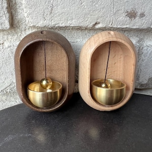 Puede incluir: Dos carillones de viento de madera con cuencos de latón y una pequeña bola dorada suspendida en su interior. Un carillón es de madera oscura, el otro de madera clara. Los carillones están colocados contra una pared de ladrillo texturizada y clara.