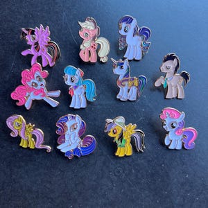 Op de afbeelding: Een verzameling emaille pins met kleurrijke cartoon pony's. Elke pin toont een ander pony-ontwerp met verschillende kleuren, waaronder roze, paars, blauw, geel en groen. De pins hebben goudkleurige metalen randen.