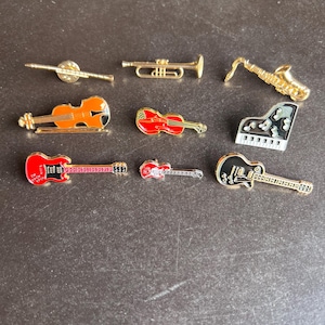 Puede incluir: Una colección de broches de instrumentos musicales, incluyendo una trompeta, un saxofón, un piano y guitarras. Los broches son dorados y rojos, con diseños detallados. Los instrumentos están dispuestos sobre una superficie oscura.