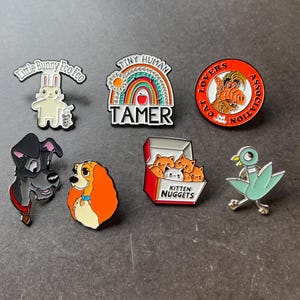 May include: A collection of enamel pins featuring various designs. Includes a white bunny pin with the text "Little Bunny Foo Foo", a rainbow pin with the text "Tiny Human Tamer", and a red pin with the text "Cat Lovers Association".