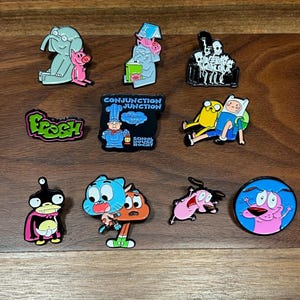 May include: A collection of enamel pins featuring cartoon characters and phrases. Designs include an elephant and pig, skeletons on a couch, the word "Fresh", characters from Adventure Time, and Courage the Cowardly Dog.