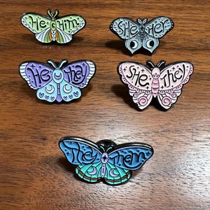May include: Five butterfly-shaped enamel pins with black outlines. Each pin features a different color scheme and pronouns: "He/Him", "She/Her", "He/They", "She/They", and "They/Them". The pins are displayed on a wooden surface.