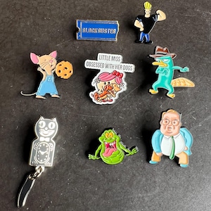 May include: A collection of enamel pins featuring cartoon characters and logos. Includes a Blockbuster pin, Johnny Bravo, Perry the Platypus, a mouse with a cookie, a cat clock, a green ghost, and a pin that says "Little Miss Obsessed With Her Dogs."