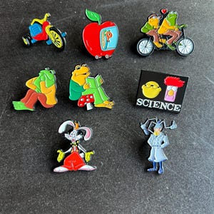 May include: A collection of eight colorful enamel pins featuring various cartoon characters and objects, including a red apple, a tricycle, a tandem bicycle, a rabbit, a detective, and a rectangular pin with the text SCIENCE and two faces.