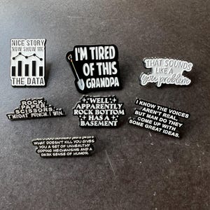 Könnte beinhalten: Eine Sammlung von schwarz-weißen Emaille-Anstecknadeln mit humorvollen Sprüchen. Die Pins zeigen Texte wie "Ich habe es satt, Opa" und "Das klingt nach einem Problem für dich". Die Pins liegen auf einer dunklen Oberfläche.