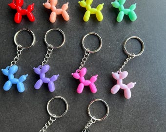 Könnte beinhalten: Eine Sammlung farbenfroher Ballonhund-Schlüsselanhänger. Jeder Schlüsselanhänger zeigt eine kleine, glänzende Hundefigur in verschiedenen Farben, darunter Rot, Rosa, Gelb, Grün, Blau, Lila und Schwarz. Die Schlüsselanhänger haben silberfarbene Metallringe und Ketten.