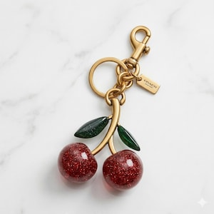 May include: A gold-toned metal keychain with a cherry design. The cherries are red with glitter, and the leaves are dark green. The keychain includes a ring, a clasp, and a small tag that says "COACH".
