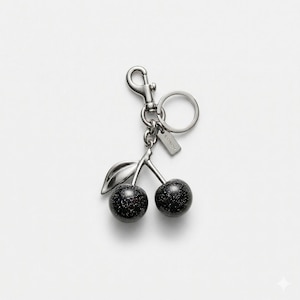 May include: A silver-toned keychain featuring a cherry design. The cherries are black with a speckled pattern, and the stem and leaf are silver. The keychain includes a clasp, a ring, and a small tag with the brand name.