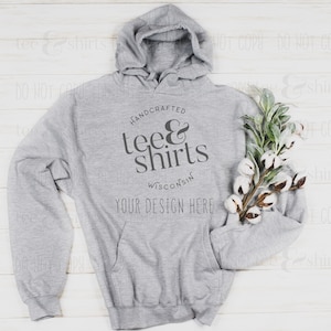 Op de afbeelding: Een grijs hoodie met de tekst "HANDCRAFTED tee & shirts WISCONSIN YOUR DESIGN HERE" gedrukt op de voorkant. De hoodie ligt plat op een wit houten oppervlak met een takje katoen.