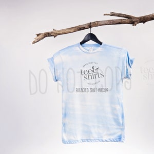 Puede incluir: Una camiseta azul claro blanqueada con un patrón tie-dye colgada de una rama. La camiseta tiene un logotipo que dice "Handcrafted Tee & Shirts Wisconsin" y "Bleached Shirt Mockup".