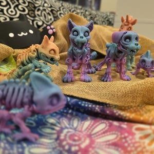 May include: A collection of colorful, skeletal animal figurines, including dogs, cats, and a chameleon. The figures are primarily blue and purple, with hints of orange and green. They are arranged on a burlap surface.