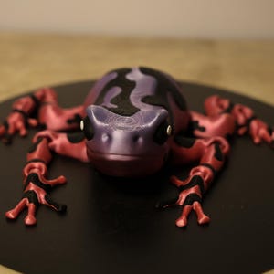 May include: A purple and black frog figurine with pink limbs. The frog is sitting on a black circular surface.