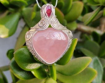May include: A silver pendant necklace with a heart-shaped rose quartz stone. The stone is set in a wire-wrapped design, with a small red gemstone at the top. The necklace is shown against a background of green leaves.