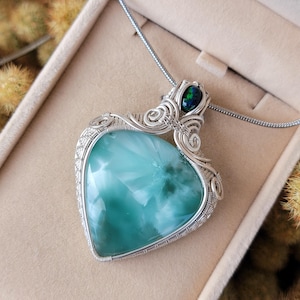 Op de afbeelding: Een hartvormige hangerketting met een lichtblauwe edelsteen, draad gewikkeld in zilver. Een kleine, donkergroene steen is boven het hart geplaatst. De ketting wordt gepresenteerd in een beige sieradendoos.