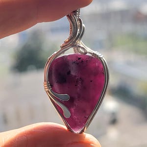 May include: A silver wire wrapped pendant featuring a purple and black gemstone. The pendant is shaped like a teardrop and has a silver bail.