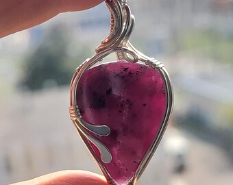 May include: A silver wire wrapped pendant featuring a purple and black gemstone. The pendant is shaped like a teardrop and has a silver bail.