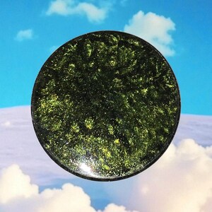 May include: A round, dark green object with a speckled, glittery texture. The object has a black border and is set against a backdrop of a blue sky with fluffy white clouds. The overall aesthetic is natural and organic.
