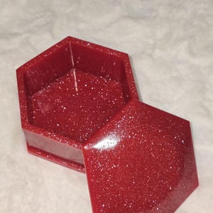 Puede incluir: Una caja de baratijas hexagonal roja con un acabado brillante. La caja está abierta, revelando el interior. La tapa está ligeramente entreabierta, mostrando el interior de la caja. La caja está hecha de un material brillante.