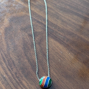 May include: Silver-toned necklace with a round pendant featuring a striped design in red, orange, blue, and green. The pendant is attached to a delicate chain, creating a simple yet colorful accessory.