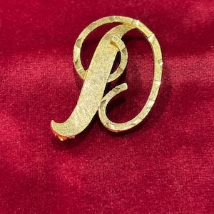 May include: A gold-coloured letter "D" brooch, set against a red velvet background. The brooch features a textured surface and a stylised design. The letter is in a cursive font, with a loop at the top and a curved bottom.