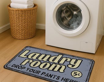 May include: A rectangular laundry room mat with the text "Laundry Room Drop Your Pants Here!" in cream-colored letters on a blue background. A washing machine and a wicker laundry basket with white towels are in the background.