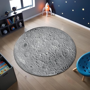 Può includere: Un tappeto rotondo con un design dettagliato della superficie lunare. Il tappeto è principalmente grigio con un aspetto strutturato, che imita i crateri e le caratteristiche della luna. È posizionato su un pavimento in legno chiaro in una stanza a tema spaziale.