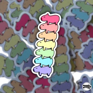 Könnte beinhalten: Ein Aufkleber mit einem Stapel von sieben bunten Capybaras, jeweils in einem anderen Pastellton, von Rosa bis Lila. Der Aufkleber hat einen weißen Rand und ist von anderen ähnlichen Aufklebern umgeben. Der Text "spoonie central co." befindet sich unten rechts.