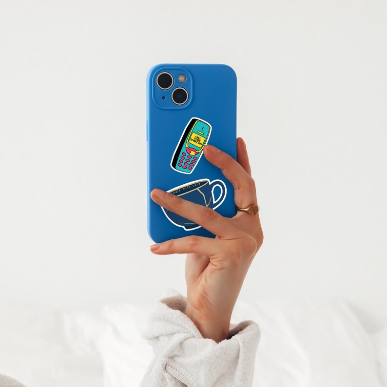 A hand wearing a white sleeve holds a blue phone with stickers of ablue and yellow 2000's style phone that says "CALL YOUR SENATORS" and coffee cup. The background is plain white, conveying simplicity.