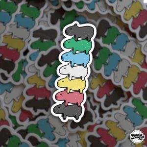May include: A stack of seven colorful capybara stickers. The capybaras are in shades of gray, green, blue, white, yellow, pink, and black. Each sticker has a white border. The Spoonie Central Co. logo is in the bottom right corner.
