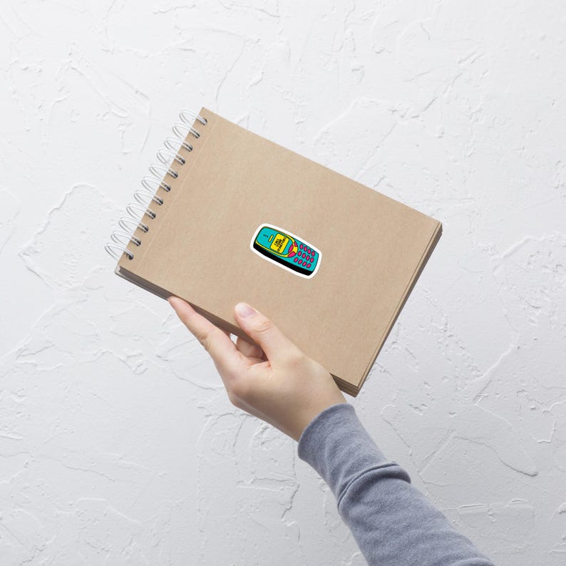 A hand holds a spiral-bound notebook with a kraft paper cover. A sticker of a blue and yellow 2000's style phone that says "CALL YOUR SENATORS" is on the cover. The background is textured white.