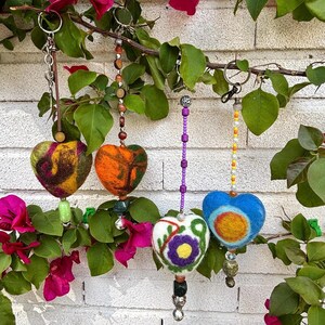 Può includere: Quattro portachiavi a forma di cuore con dettagli di perline sono appesi contro un muro di mattoni bianchi, circondati da foglie verdi e fiori rosa. I cuori sono infeltriti in vari colori e motivi, tra cui arancione, blu e bianco con disegni floreali.