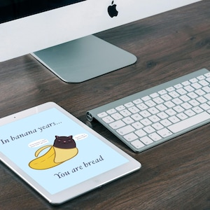 May include: A white tablet displaying a light blue background with a cartoon illustration of a black cat inside a yellow banana peel. The text reads In banana years... You are bread, with two small speech bubbles containing illegible text.