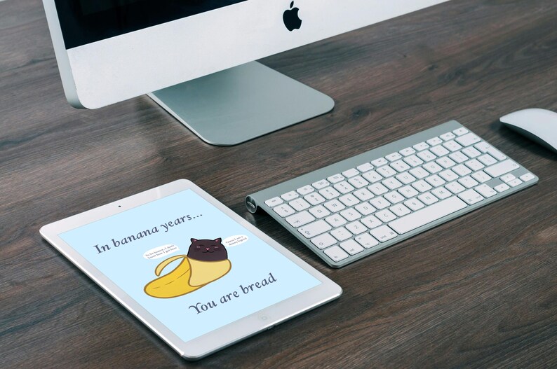 May include: A white tablet displaying a light blue background with a cartoon illustration of a black cat inside a yellow banana peel. The text reads In banana years... You are bread, with two small speech bubbles containing illegible text.