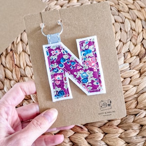 May include: A fabric letter N keyring featuring a purple floral pattern with blue and pink flowers, outlined in white felt. The letter is attached to a silver metal ring with a light blue ribbon and mounted on a brown card with the text DOTTY & DARLING.