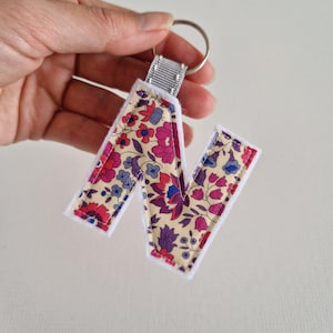 May include: A fabric letter "N" keyring with a silver key ring. The letter features a cream-coloured floral pattern with pink, purple, and blue flowers. It has white fabric edging and a grey ribbon loop for attachment.