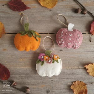May include: Three felt pumpkin ornaments with hanging loops. One is orange with a green stem, one is dusty rose with white embroidered vine details, and one is white with a cluster of small felt flowers in pink, orange, and white.