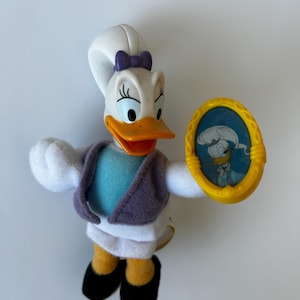 Può includere: Un'anatra di peluche bianca con un fiocco viola, indossa una giacca blu e viola e una gonna bianca, tiene un telaio giallo con un'immagine di un'anatra all'interno.
