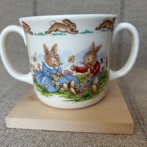May include: Cream-colored ceramic mug with two handles, featuring illustrations of rabbits in a garden setting. The design includes a rabbit couple, flowers, and running rabbits around the rim. The mug sits on a small wooden square.