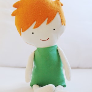 May include: A handmade plush toy doll with orange hair, a white face, and a green shirt. The doll is sitting with its legs crossed.
