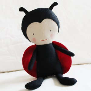 Puede incluir: Un juguete de peluche en forma de mariquita, hecho de tela negra y roja. La mariquita tiene una cara sonriente con ojos negros y mejillas rosadas.