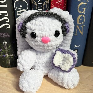 May include: A white crocheted bunny with a gray headband and purple accents. The bunny is holding a purple and white crocheted flower.