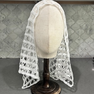 May include: A white crochet head covering featuring an openwork grid pattern. The fabric is draped over a beige mannequin head mounted on a dark brown wooden stand. The lace-like textile displays a repeating geometric design throughout the piece.