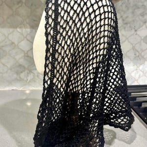 Puede incluir: Un cubrecabeza negro con un patrón de ganchillo abierto similar a una red. La tela está construida con un diseño de malla repetitivo en forma de diamante, creando una textura ligera y aireada que cae desde la parte superior del soporte.