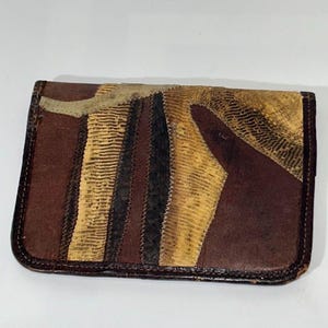 May include: A rectangular brown leather wallet with a patchwork design. The wallet features various textures and colors, including dark brown, tan, and black, with a stitched border. The design is a mix of geometric shapes and patterns.