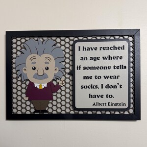 May include: A black framed wall art piece with a cartoon illustration of Albert Einstein. The quote "I have reached an age where if someone tells me to wear socks, I don't have to." is printed on a white background with a gray border. The quote is attributed to Albert Einstein.