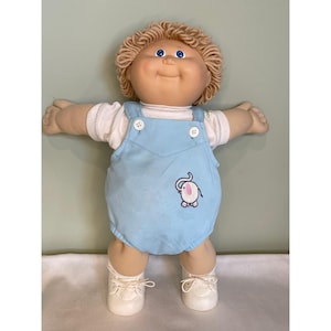 Op de afbeelding: Een vintage Cabbage Patch Kid pop met blond, krullend haar en blauwe ogen. De pop draagt een wit shirt onder een lichtblauwe overall met een geborduurd olifantenontwerp. Witte schoenen maken de outfit compleet.
