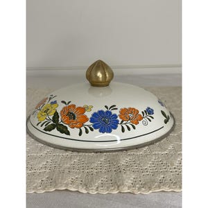 Puede incluir: Una tapa de esmalte blanco con un pomo dorado y un diseño floral en amarillo, naranja y azul. La tapa tiene un borde plateado y probablemente sea para una olla o sartén. El patrón floral está dispuesto alrededor del borde de la tapa.