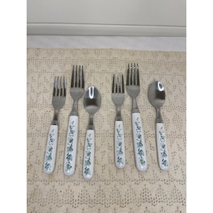 May include: A set of six pieces of cutlery including four stainless steel forks and two stainless steel spoons. Each piece features a white handle decorated with a repeating green and grey floral pattern along the length of the handle.