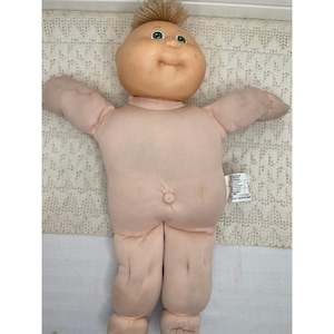 May include: A soft-bodied doll with a round, light-toned vinyl head, green eyes, and short, light brown yarn hair. The doll has a pale, fabric-covered torso, arms, and legs, featuring a small belly button and a white fabric tag on the side.